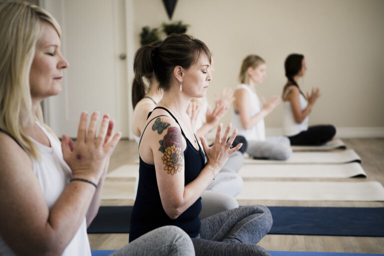 Women seated in meditation during a restorative yoga class at The Rooted & Rising, supporting healing from chronic stress symptoms through mindful movement.